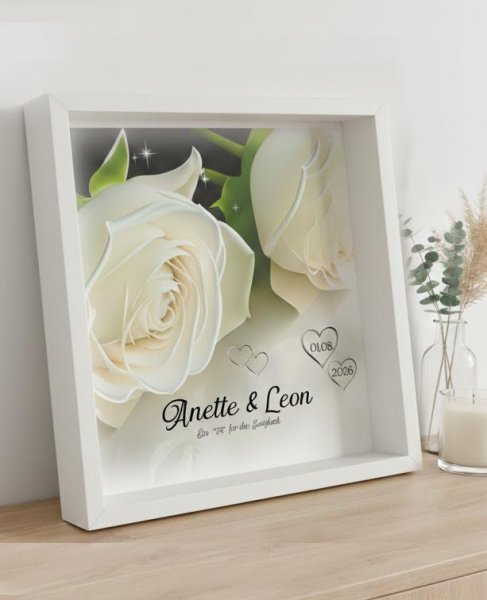 Personalisiertes Hochzeitsgeschenk-Set: Eine quadratische Wanduhr und ein passendes Dekobild in tiefen weißen 25x25 cm Objektrahmen auf einem Sideboard. Die Uhr zeigt ein edles Ring-Motiv mit Namen und Datum, das Bild eine weiße Rose mit Personalisierung.